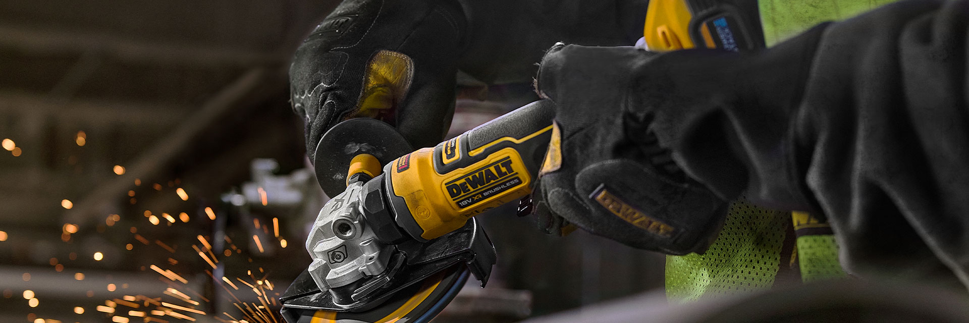 A person using a DEWALT cordless angle grinder to cut or grind a large metal pipe, with visible sparks flying from the grinding area. The person is wearing protective gloves and a high-visibility vest.
