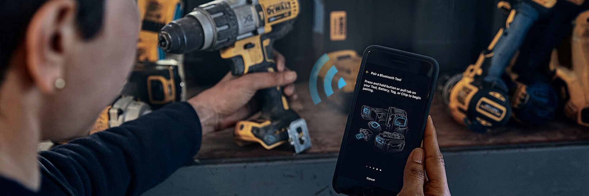 A person uses a smartphone to connect wirelessly with a DEWALT power drill on a workbench, with other DEWALT tools visible in the background.